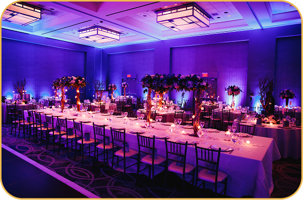 Elegant banquet hall with long tables, purple lighting, and floral centerpieces